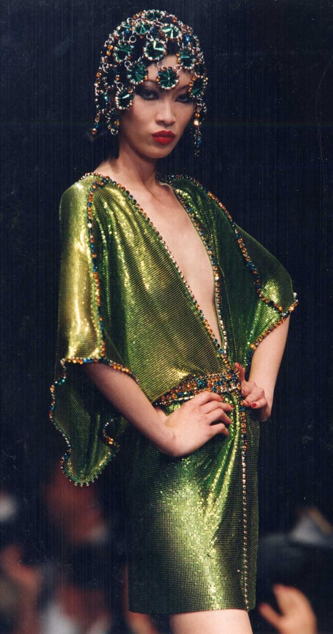 Model wearing a sparkling green dress with jeweled details and an elaborate headpiece on a runway.