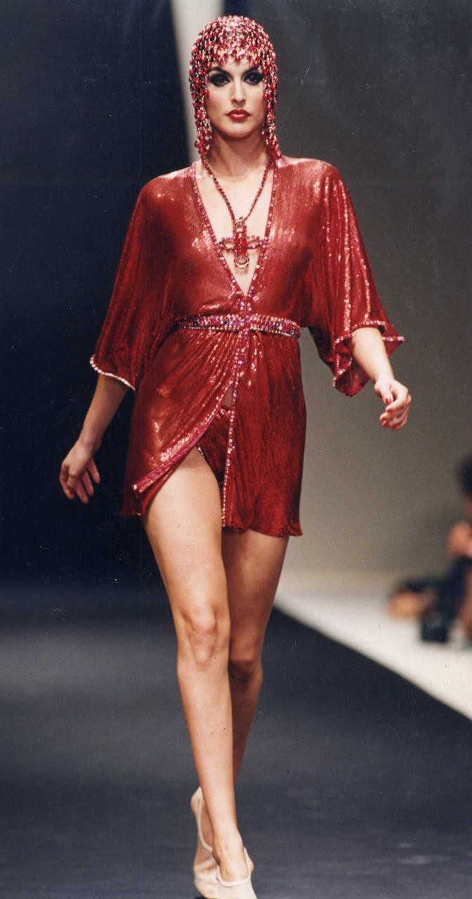 Model walking the runway in a sparkling red outfit with a beaded headpiece during a fashion show.