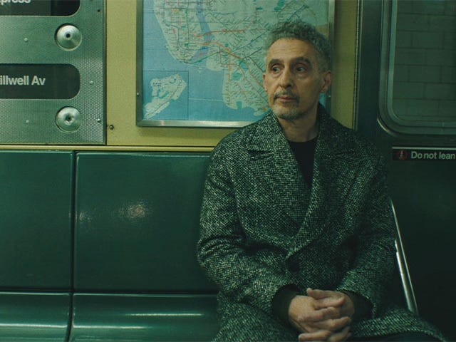 A man sitting alone on a subway bench, wearing a grey coat, with a map and signs in the background.