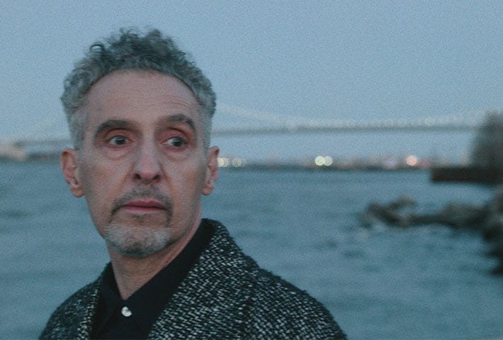 A man with curly gray hair standing by the water, looking thoughtfully into the distance with a bridge in the background.