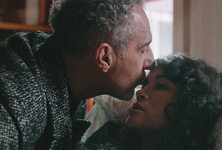 A man kisses a woman gently while she gazes at him, conveying intimacy and emotion in a cozy setting.