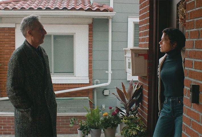 A man and woman engage in conversation at a doorway, surrounded by potted plants and a cozy residential setting.