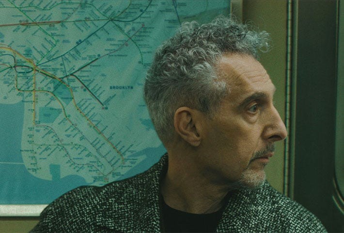 Man with curly gray hair sitting in a subway, looking toward a map of the Brooklyn area in the background.