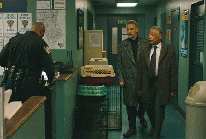 Two men walking in a police station hallway, passing a desk with an officer and various items in the background.