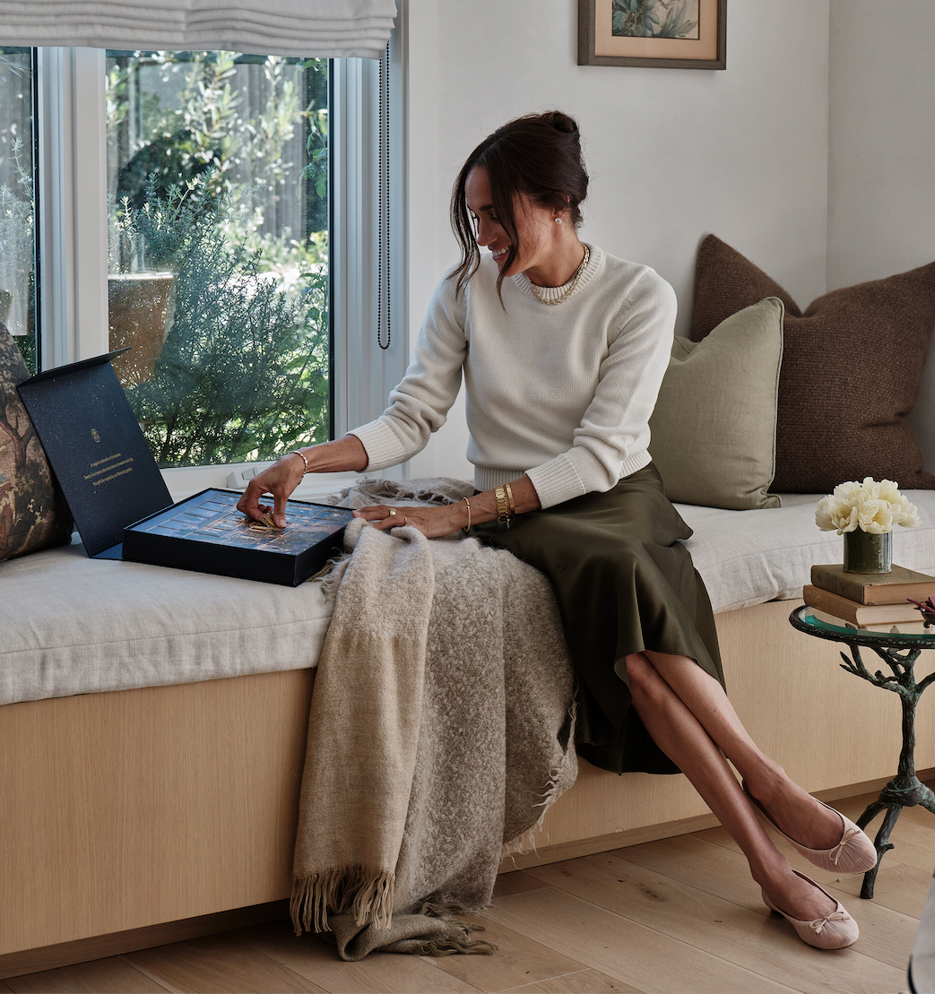 A woman sitting on a bench, examining a box while wrapped in a blanket, with plants and furnishings in a bright room.
