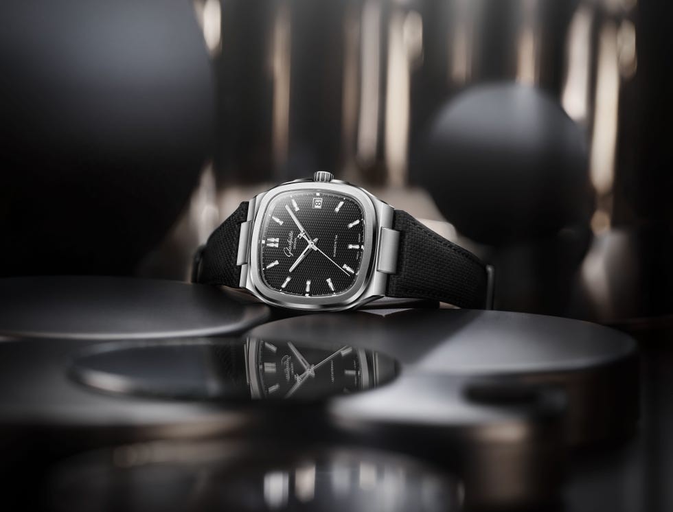 A stylish black watch with a square face and a textured strap, resting on a reflective surface.