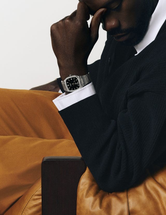 A close-up of a person with a stylish watch resting their chin on their hand while seated on a brown leather chair.