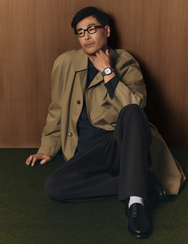 Man in a stylish coat and watch, seated against a wooden backdrop with green flooring.