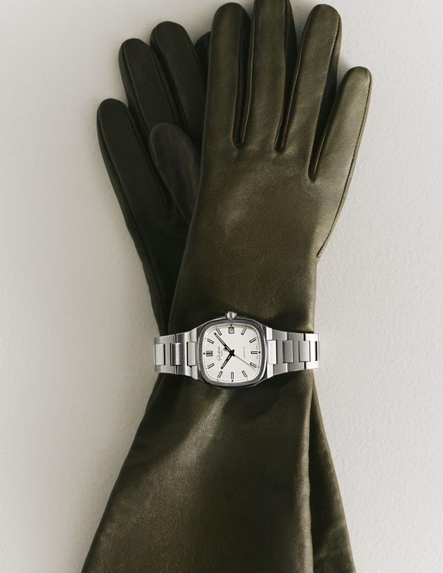 Silver watch resting on green leather gloves against a neutral background.