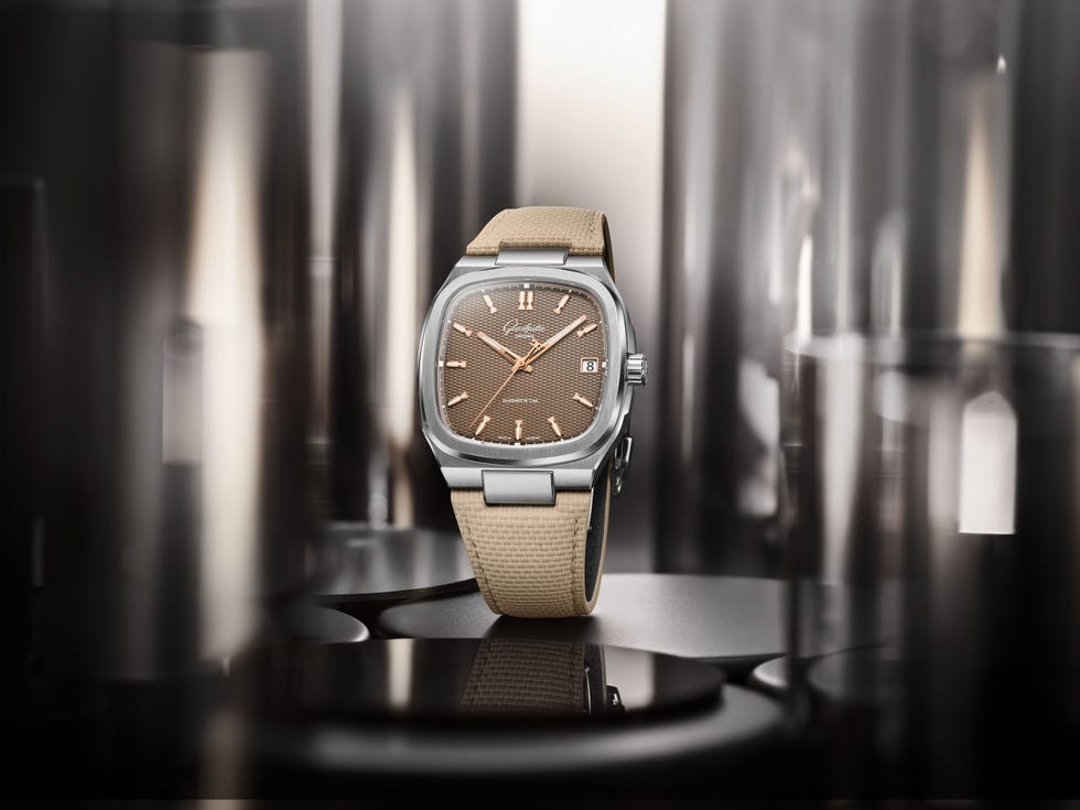 A stylish brown watch with a silver case and beige strap, set against a blurred industrial background.
