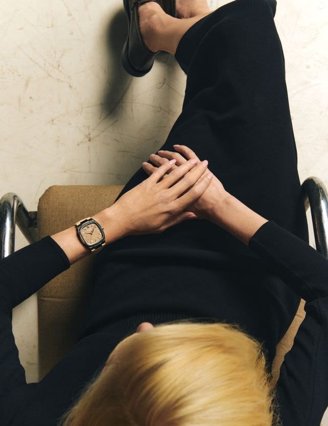 A person seated with hands clasped, wearing a black outfit and a stylish watch, with blonde hair visible.