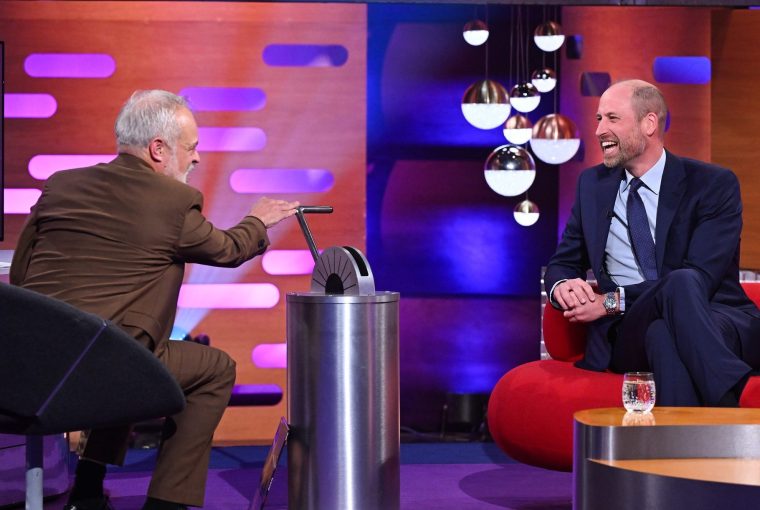 Two men laughing during a talk show, one gesturing toward a metallic device on a table.