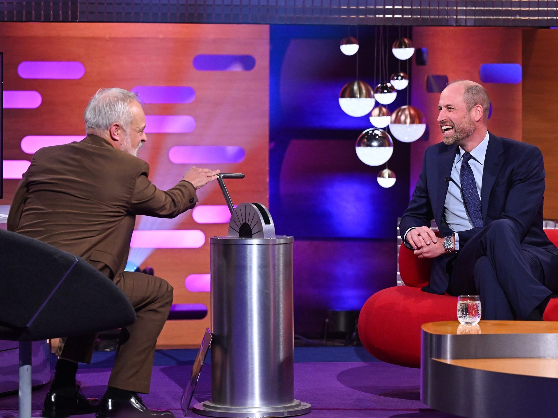Two men laughing during a talk show, one gesturing toward a metallic device on a table.