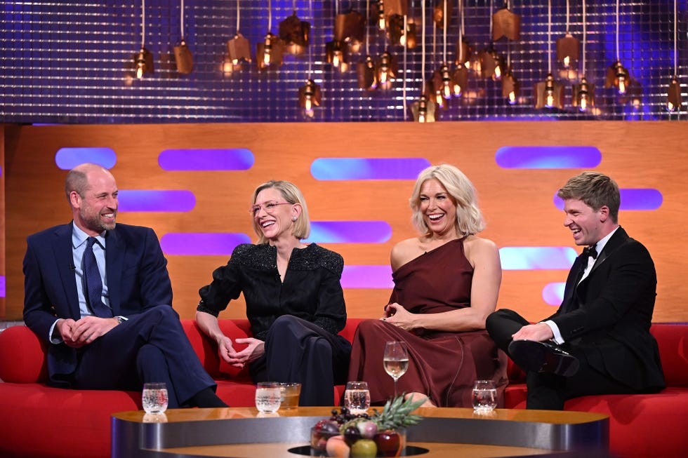 Four guests laughing and chatting on a talk show set with colorful lighting and a cozy red sofa.