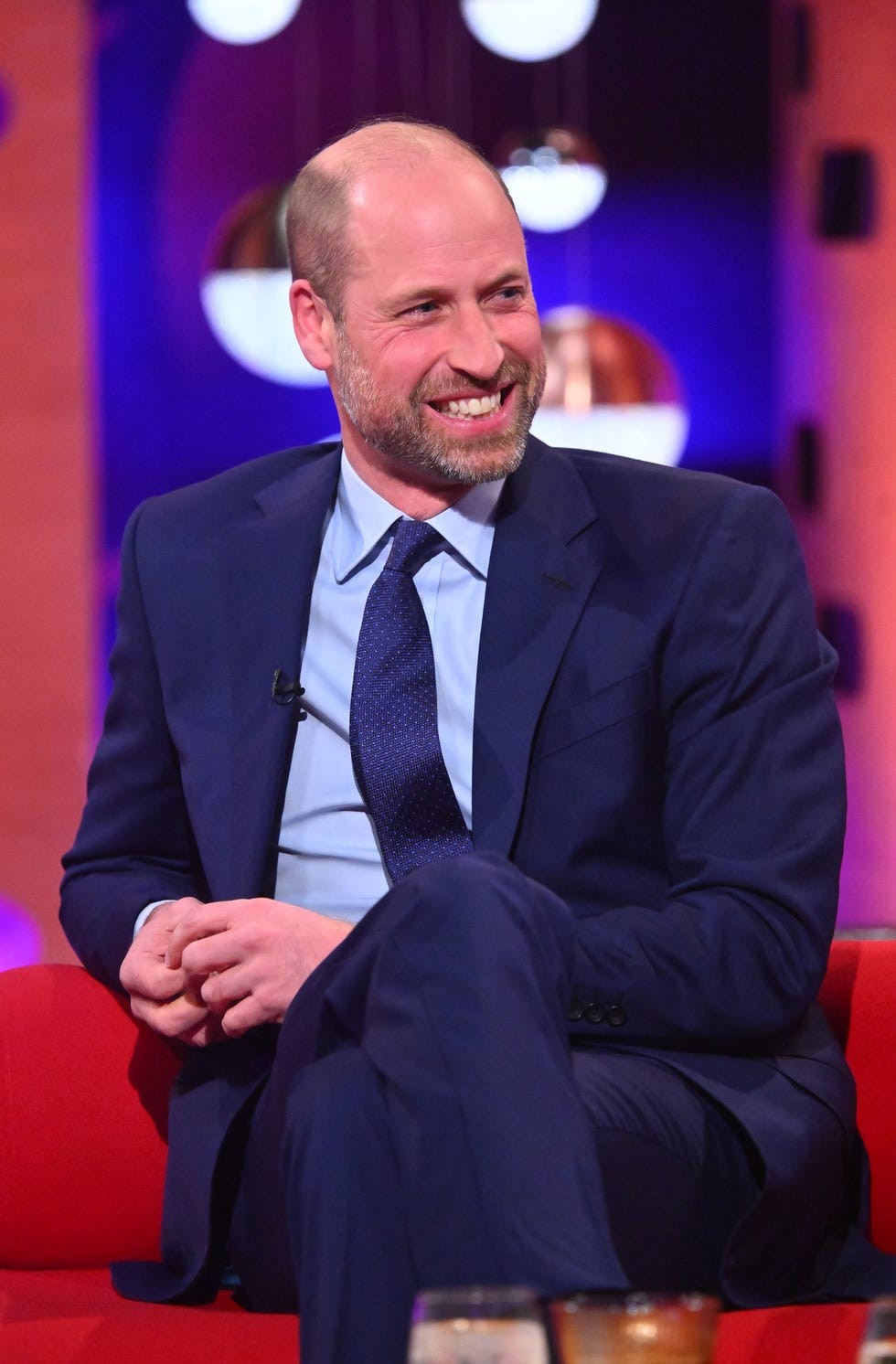 Smiling man in a suit sitting on a red couch, engaged in conversation on a talk show set.
