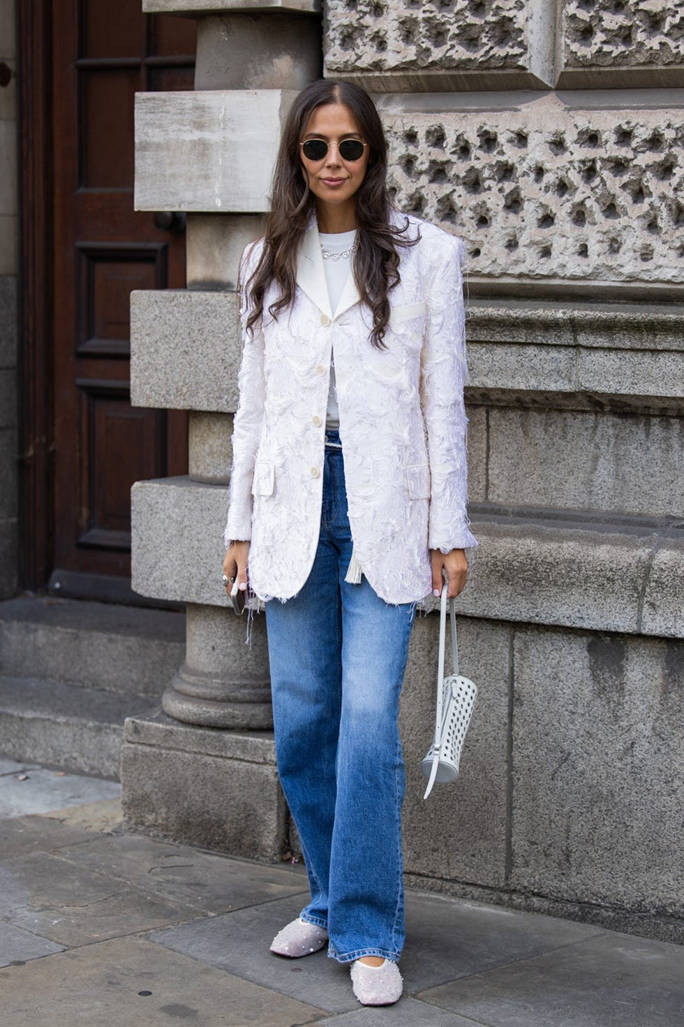 Woman wearing a textured white blazer, sunglasses, and flared jeans, standing against a stone wall.