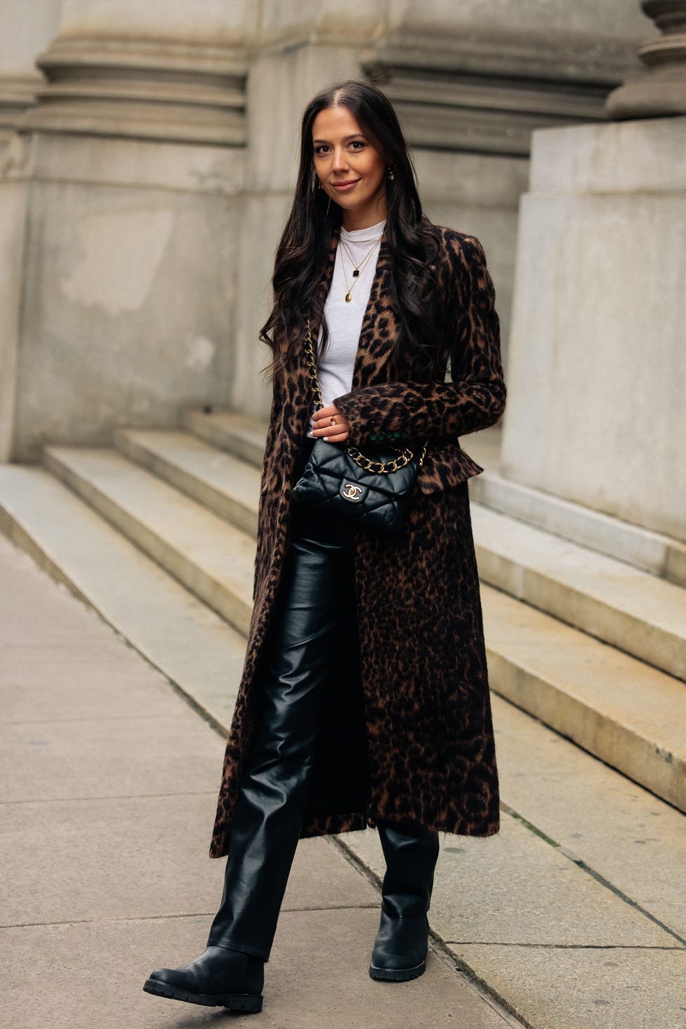 A woman wearing a leopard print coat and leather pants stands on steps, holding a black purse with a gold chain.