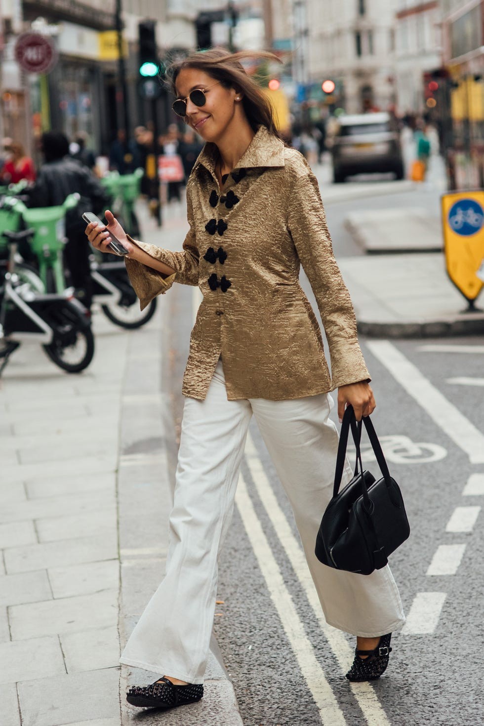 Young woman walking on a city street in a gold jacket and white pants, holding a phone and a black handbag.