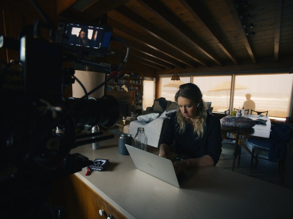 A woman working on a laptop in a cozy, sunlit room with a camera setup in the foreground.
