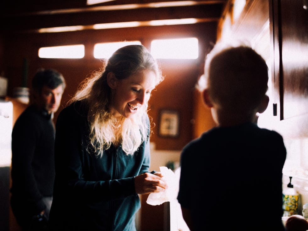A woman joyfully interacts with a small child while another person observes in a brightly lit kitchen.