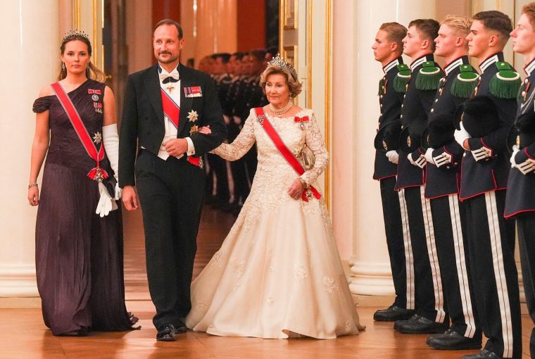 Three elegantly dressed individuals walk past a line of soldiers in formal attire during a ceremonial event.