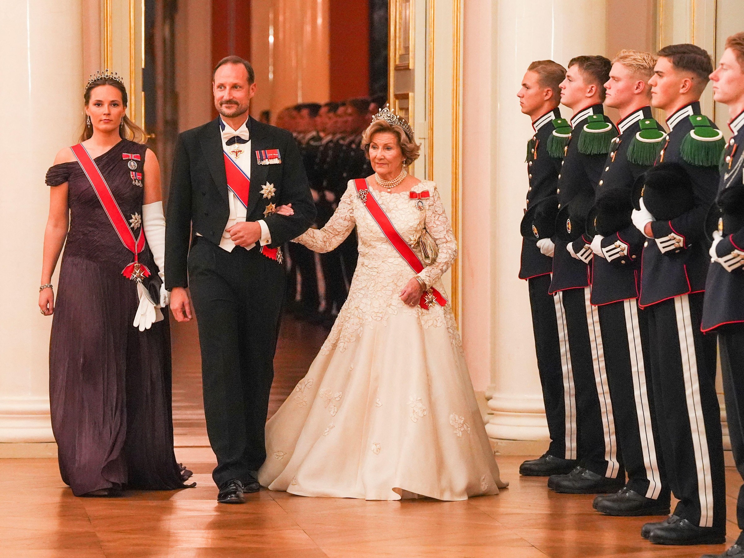 Three elegantly dressed individuals walk past a line of soldiers in formal attire during a ceremonial event.