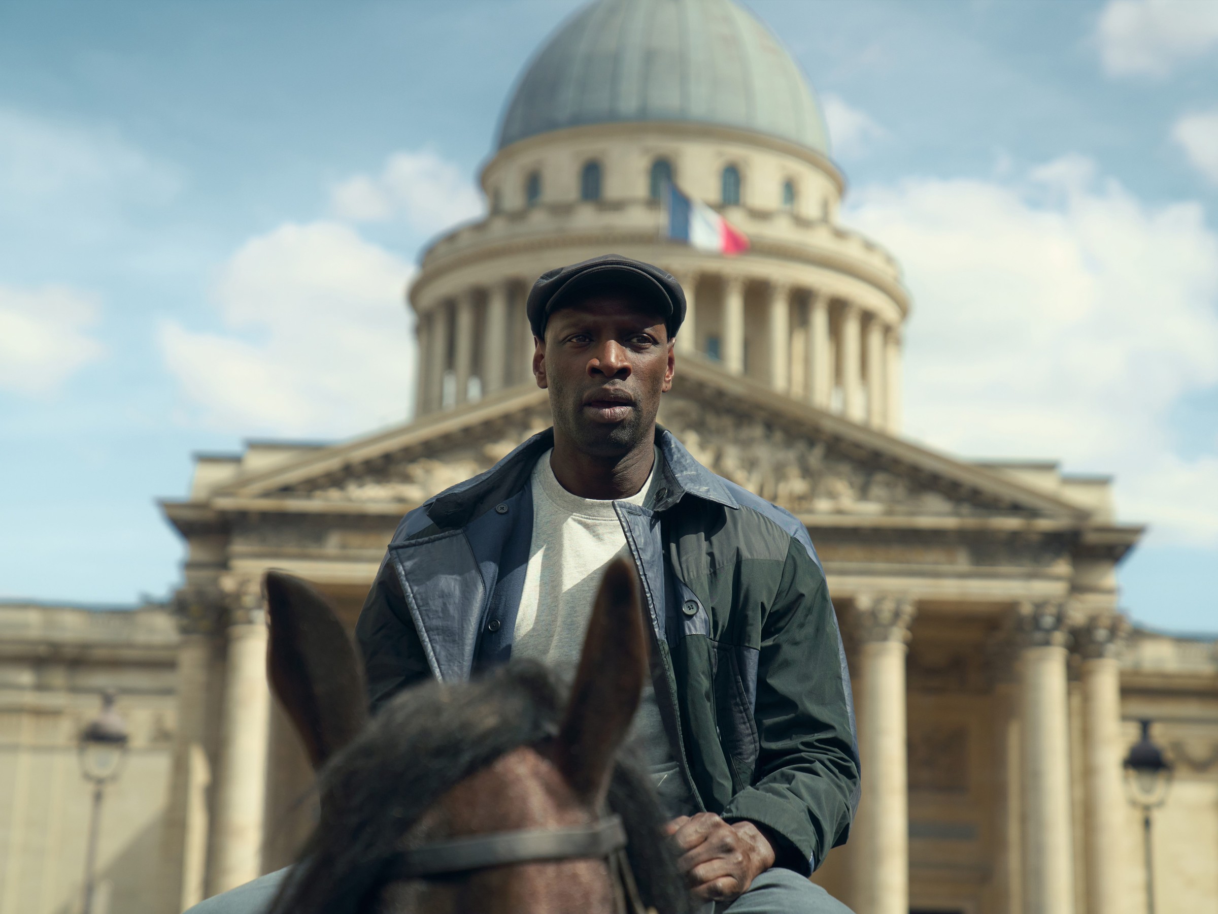 A man on a horse in front of a historic building with a dome and French flags in a clear sky.