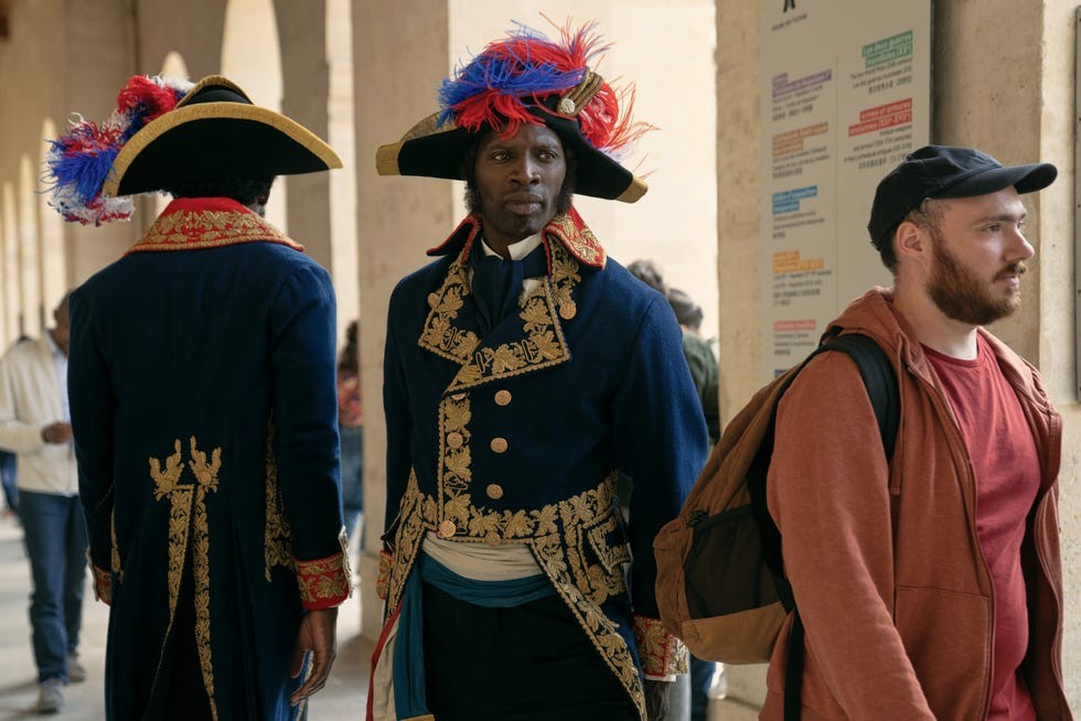 Two individuals in historical costumes, one facing forward and the other turned away, in a gallery setting.