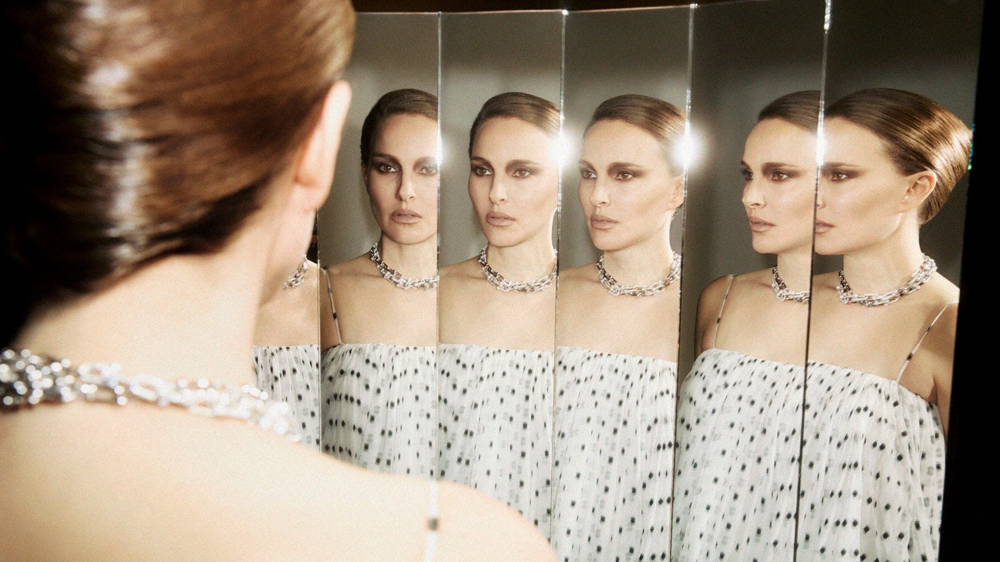 A woman in a patterned dress and necklace gazes into multiple mirrors, creating reflections of herself.