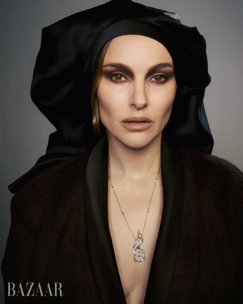 A woman in a black headscarf and robe, wearing a sparkling necklace, gazes directly at the camera.