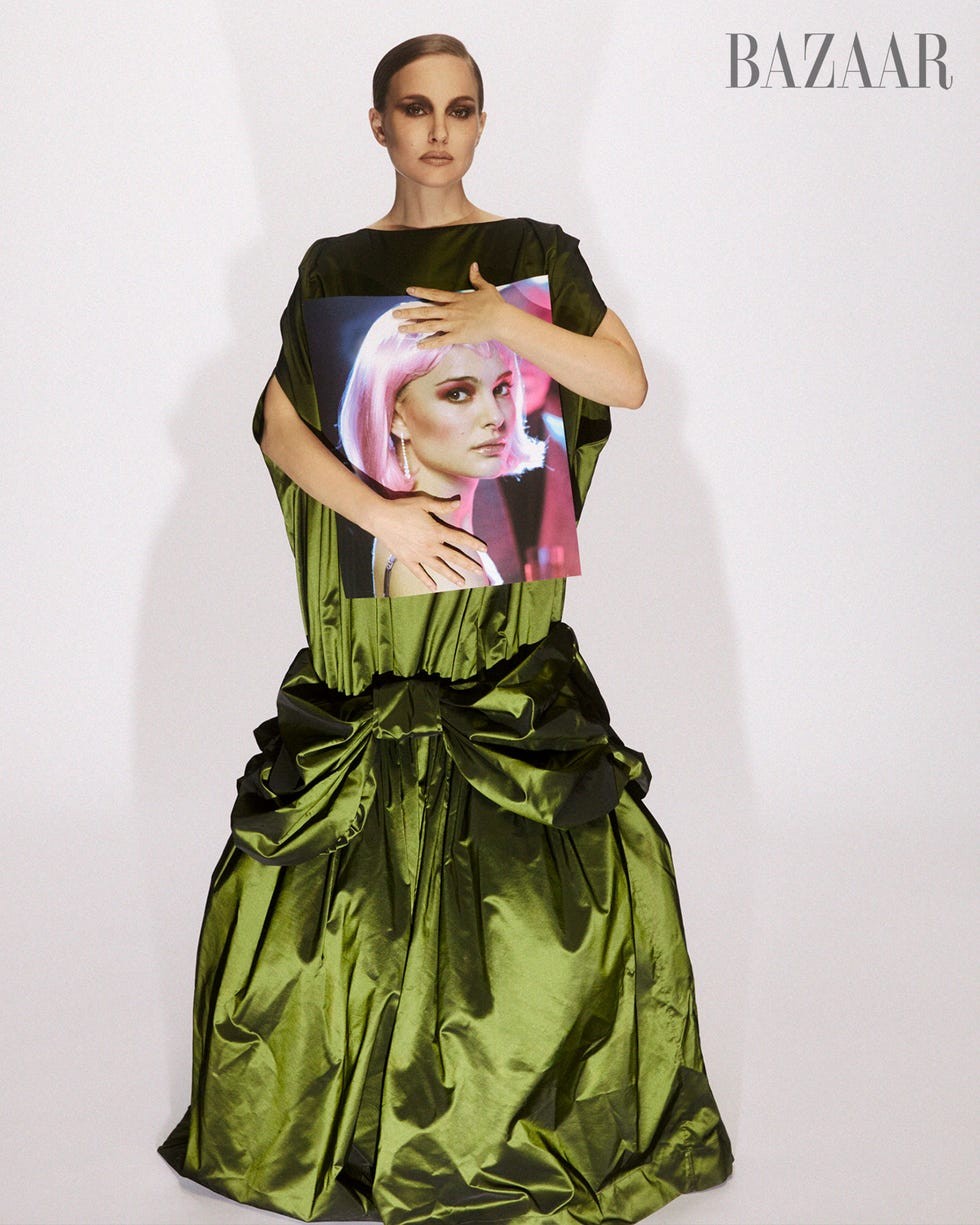 Model wearing a dramatic green gown, holding a portrait of a woman with pink hair, with a minimalist background.