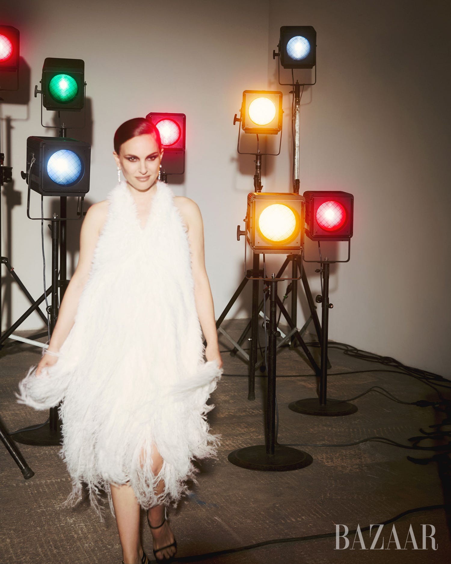A model in a white feathered dress walks under colorful studio lights.