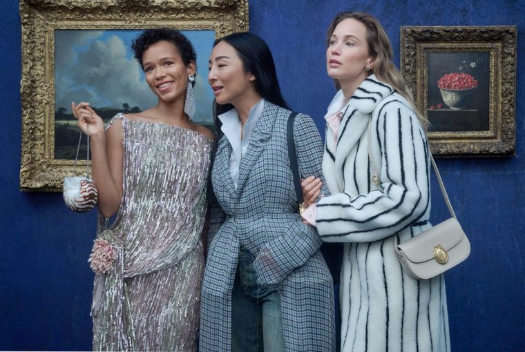 Three women in stylish attire posing together in front of a blue wall and framed artwork.
