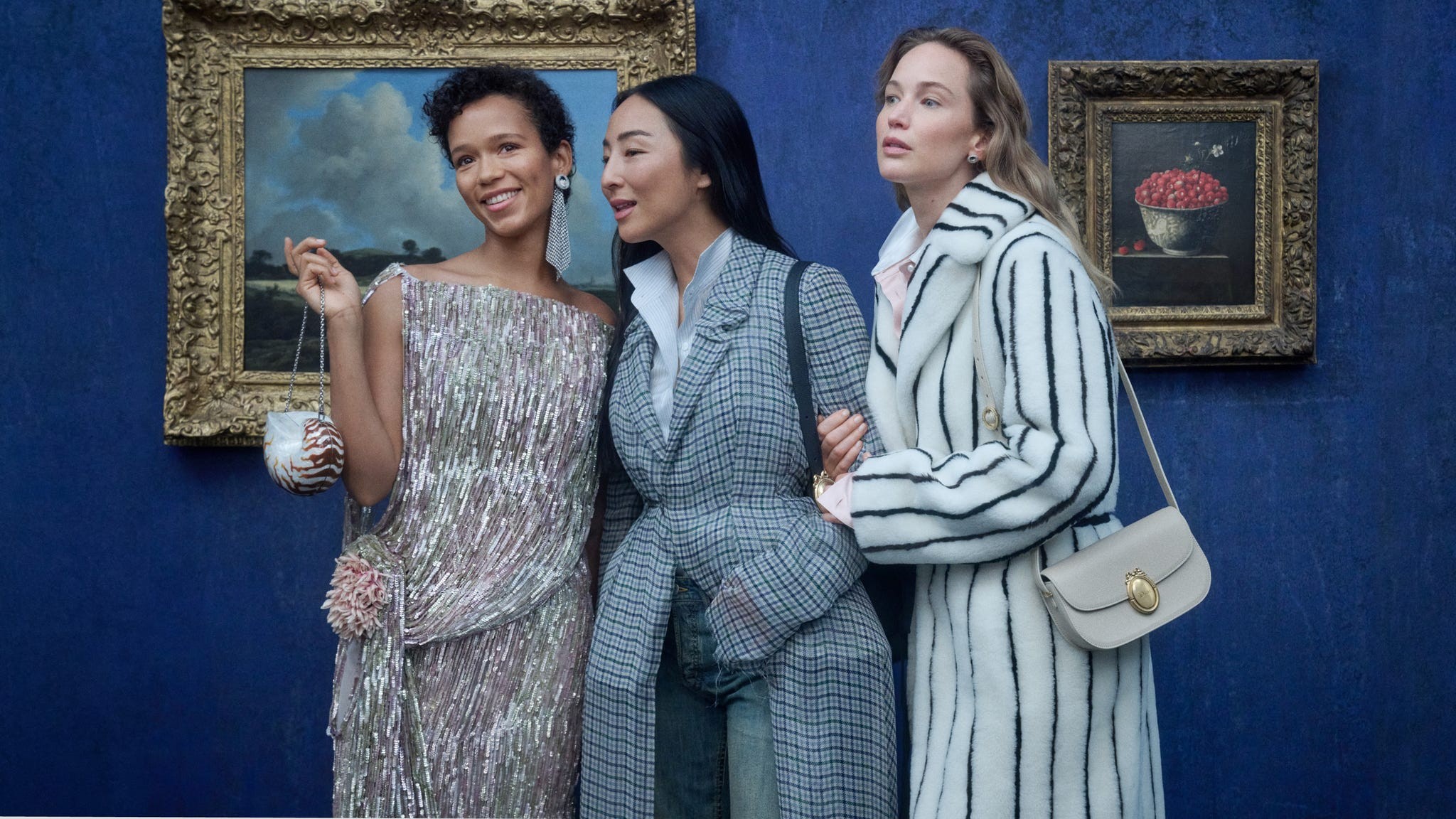 Three women in stylish attire posing together in front of a blue wall and framed artwork.