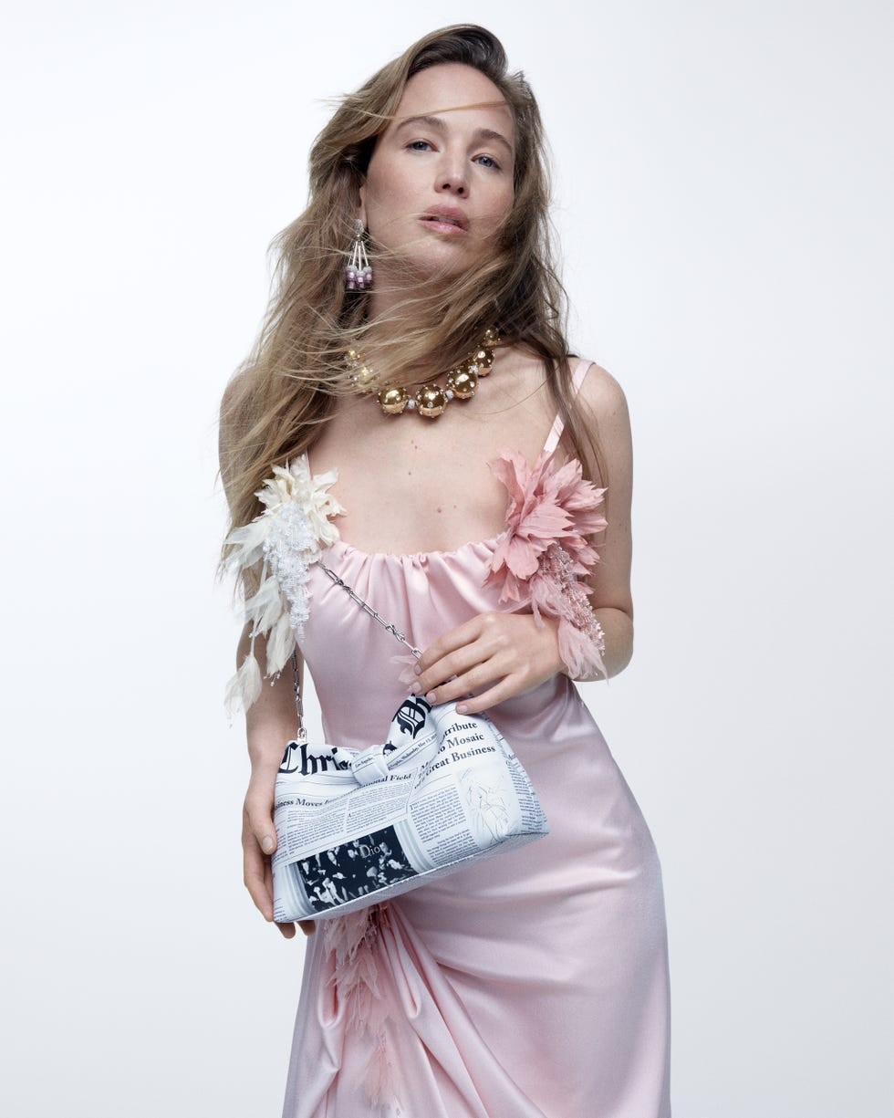 Model in a pink dress with floral accents, holding a newspaper-print handbag, with flowing hair and statement jewelry.