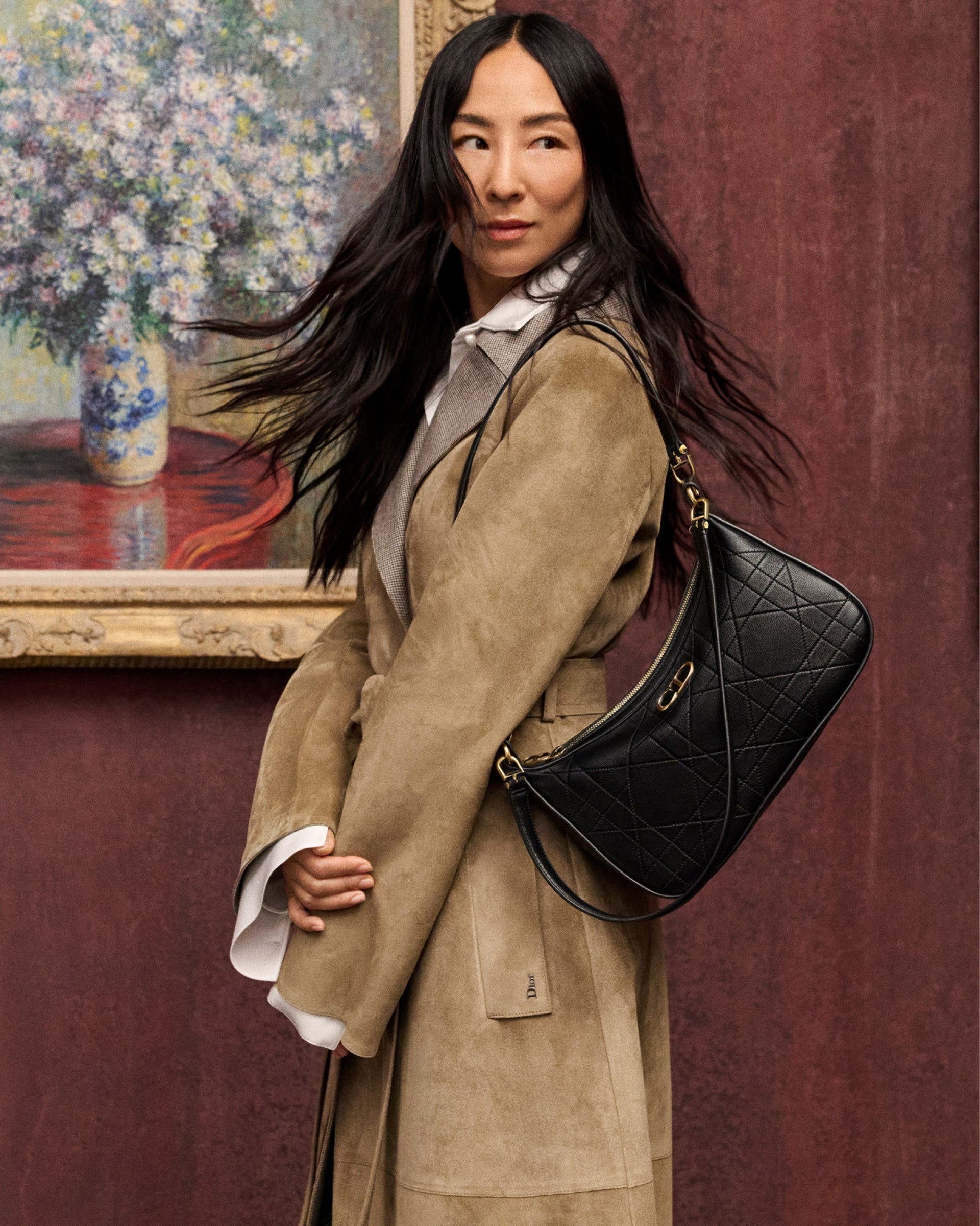 person wearing a tan coat with a black handbag, standing in front of a floral painting.
