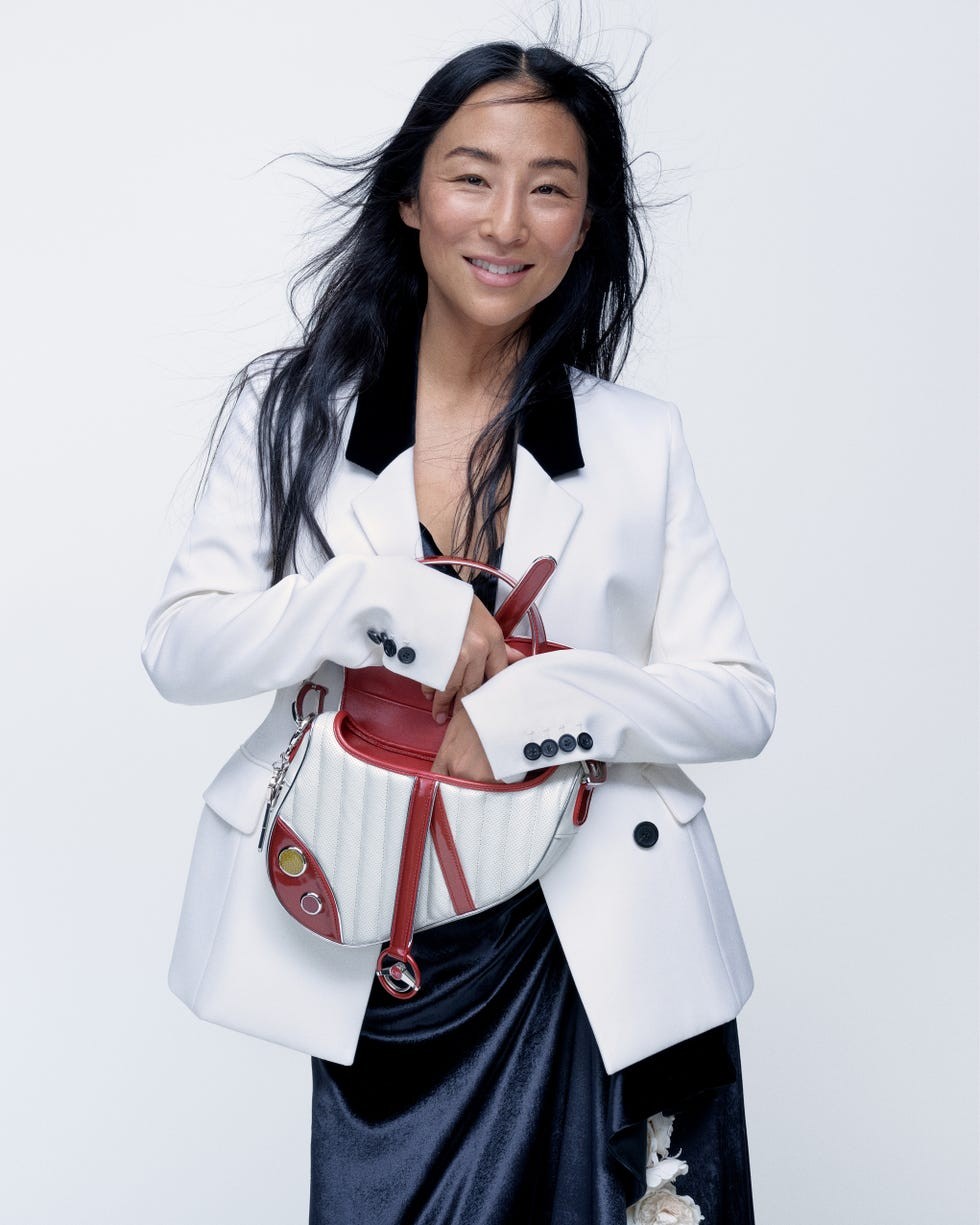 A smiling person in a white blazer holds a stylish handbag against a light background.