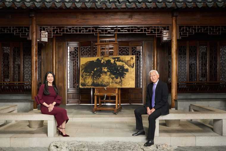 Two individuals sitting on stone benches in a traditional Chinese interior with an easel and painting in the background.