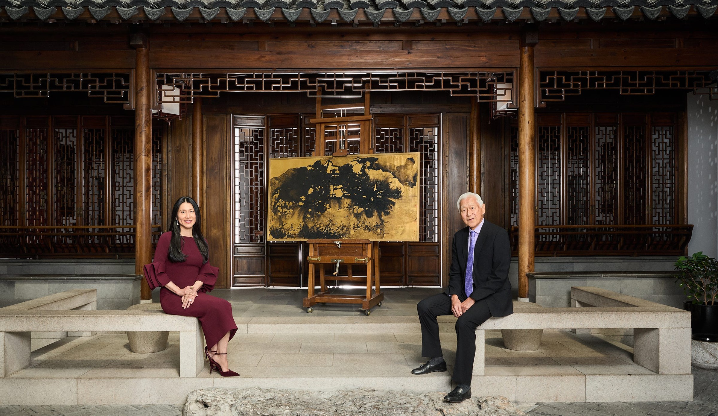 Two individuals sitting on stone benches in a traditional Chinese interior with an easel and painting in the background.
