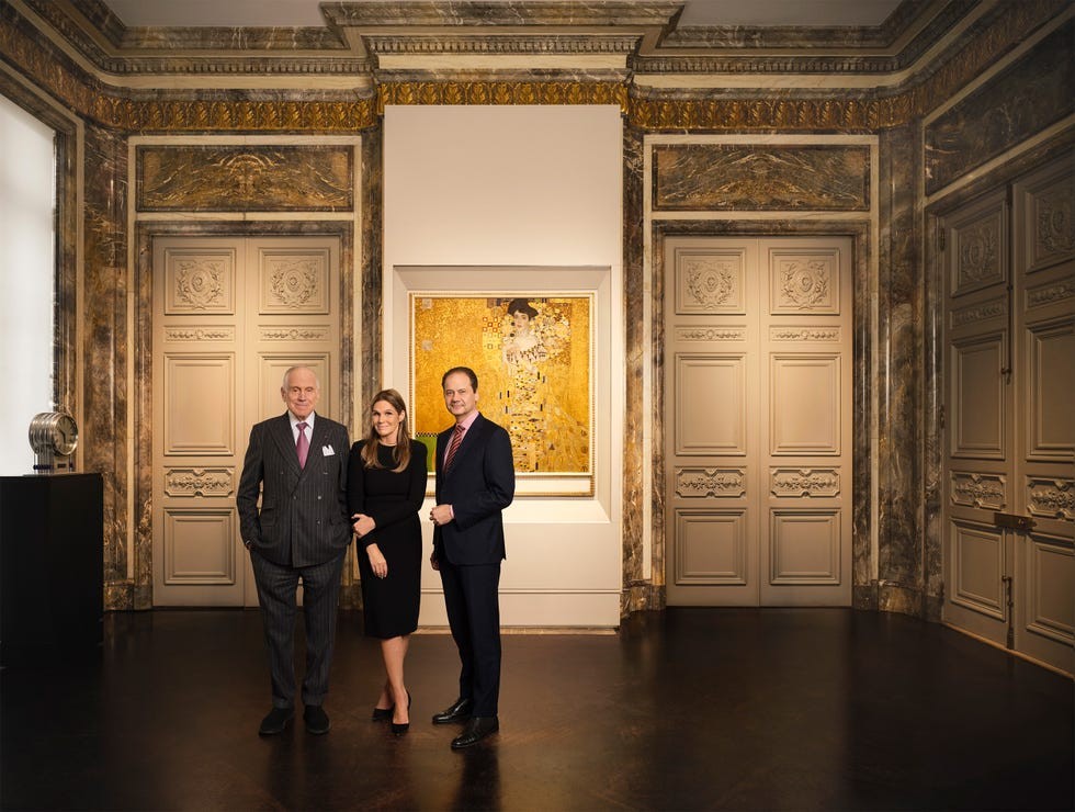 Three individuals posing in an elegant gallery with ornate decor and a prominent artwork in the background.