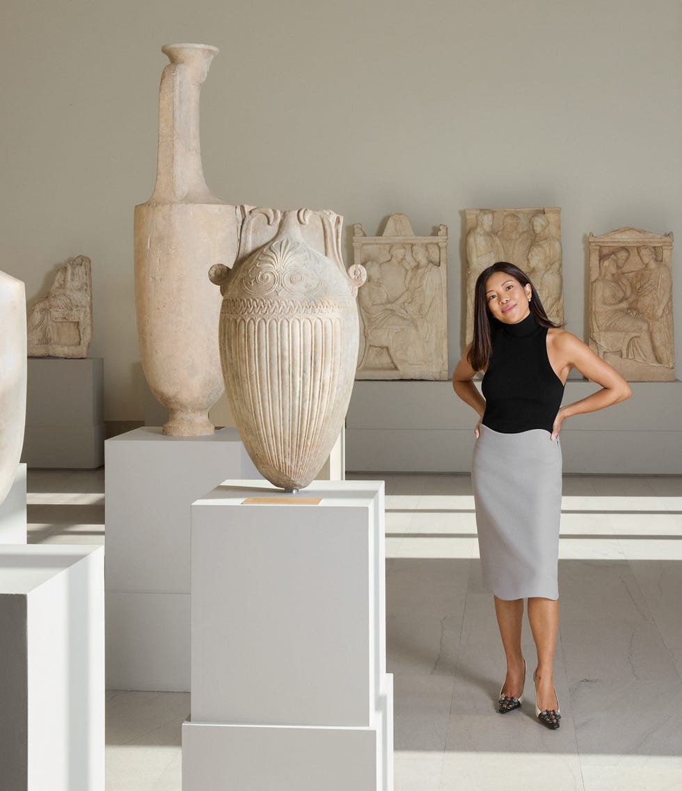 Woman in a black top and gray skirt poses beside ancient pottery in a well-lit museum gallery.