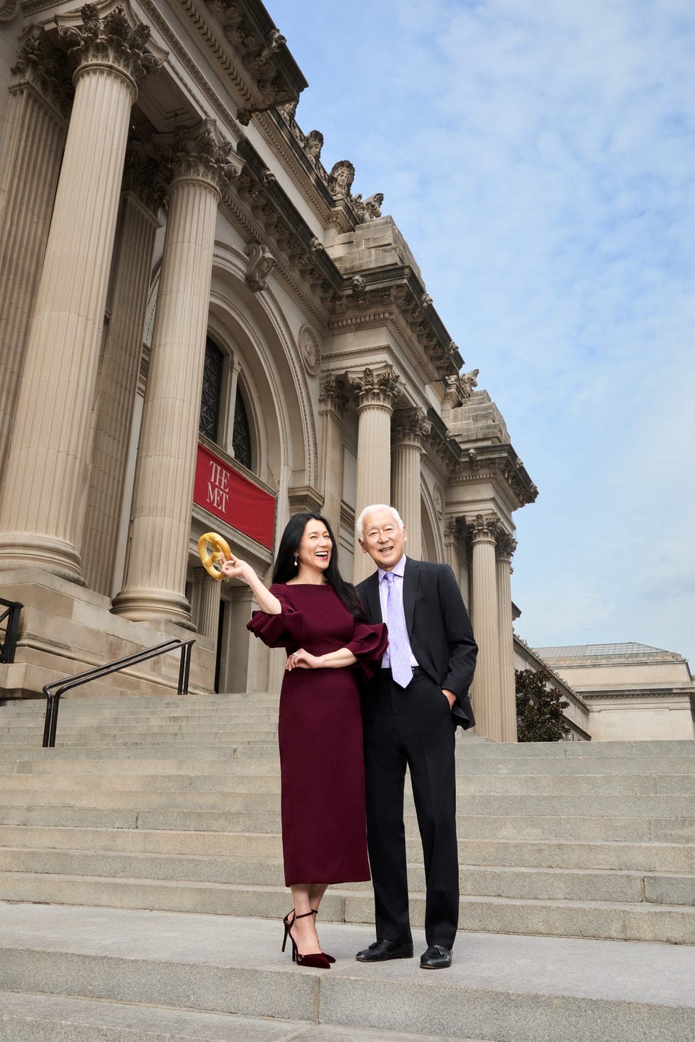 Two smiling individuals pose on the steps of a grand building, with one holding a golden object and wearing elegant attire.