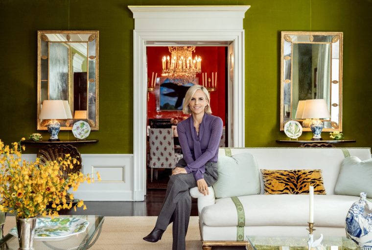 Woman sitting on a sofa in a stylish green living room with decorative mirrors and floral arrangements.