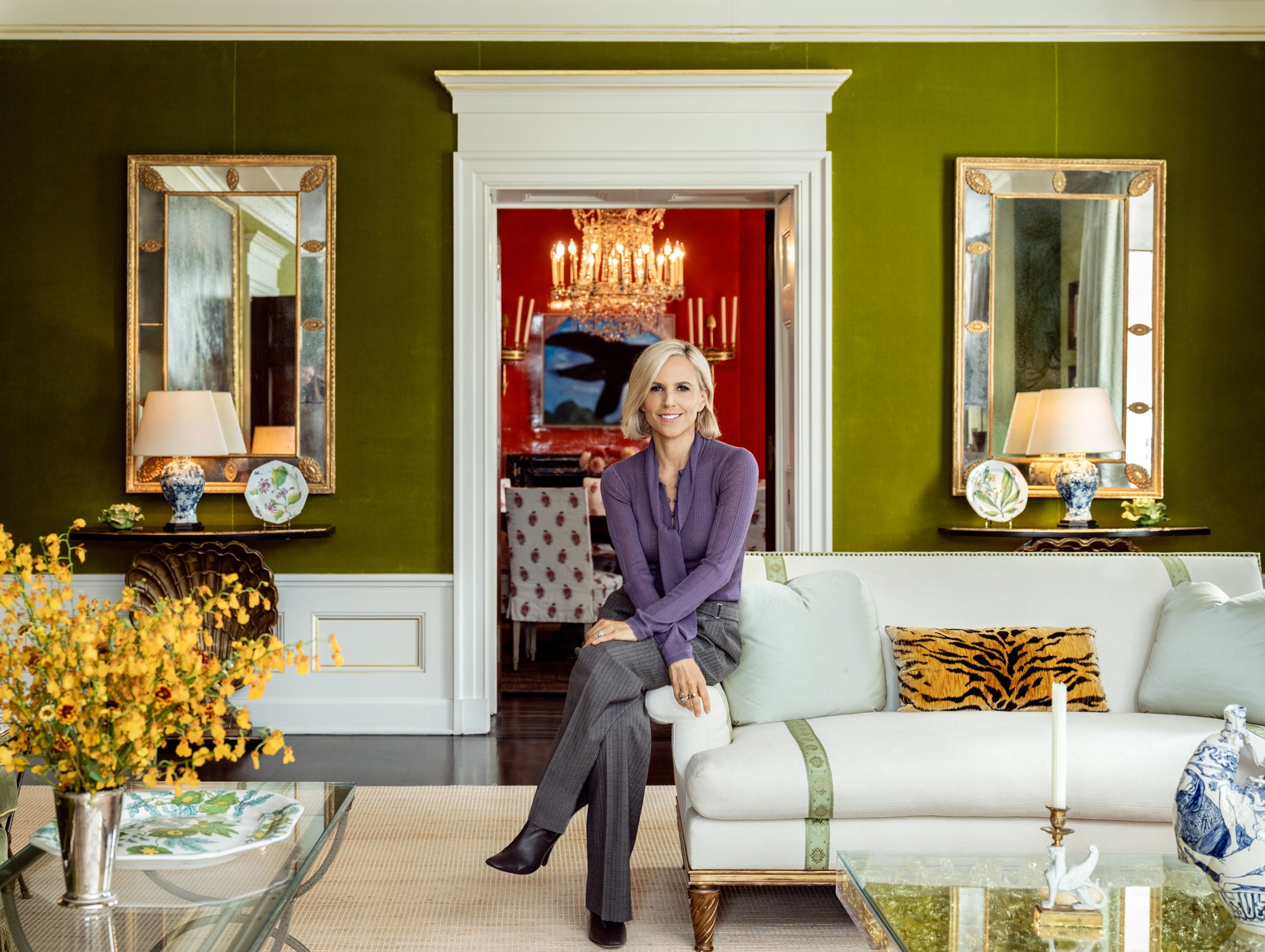 Woman sitting on a sofa in a stylish green living room with decorative mirrors and floral arrangements.