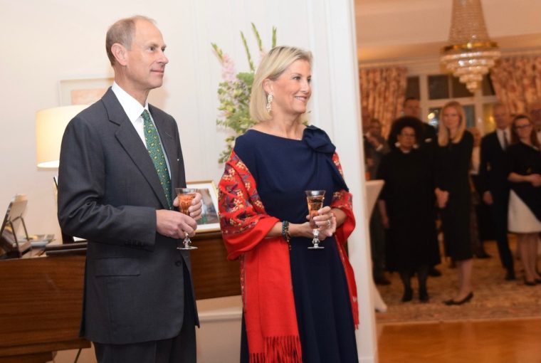 Two elegantly dressed individuals holding drinks at a formal event, with guests in the background and a piano nearby.