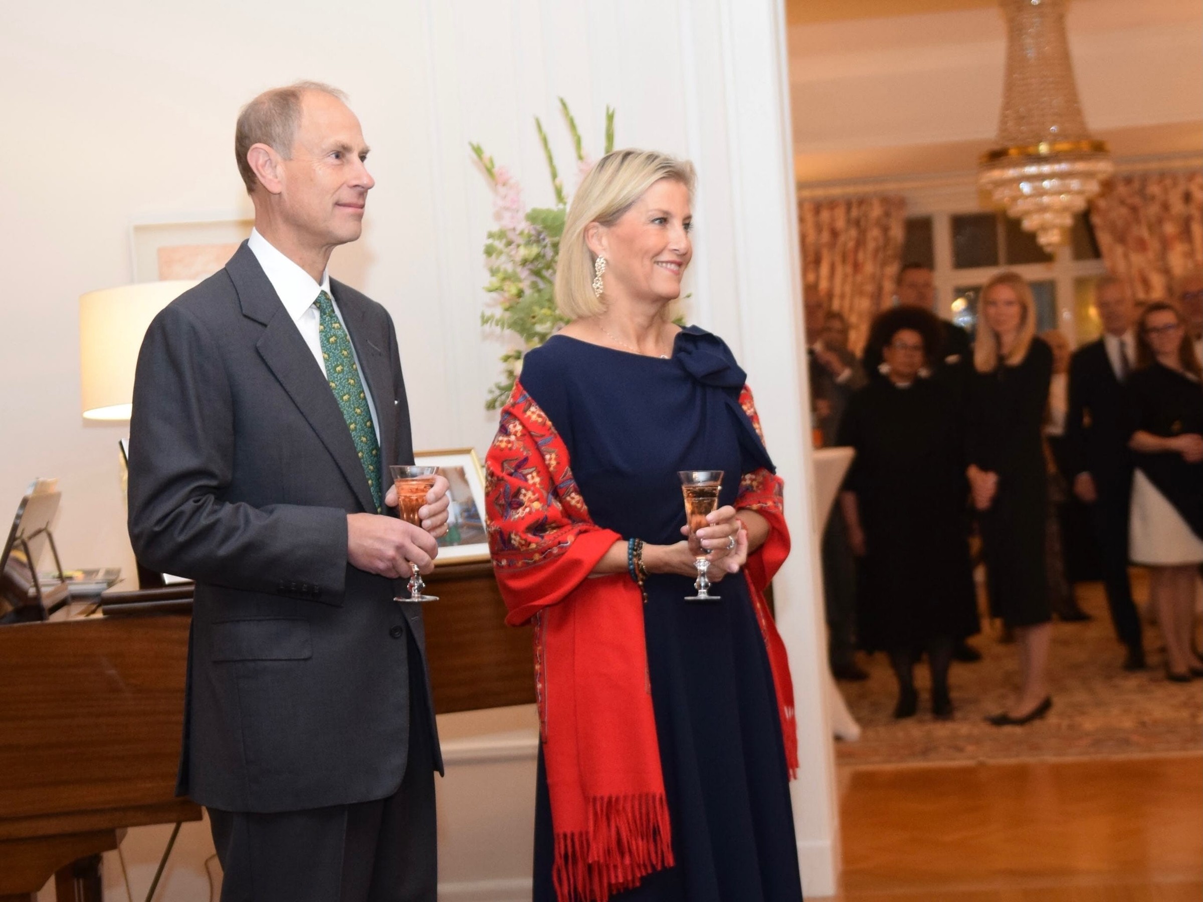 Two elegantly dressed individuals holding drinks at a formal event, with guests in the background and a piano nearby.