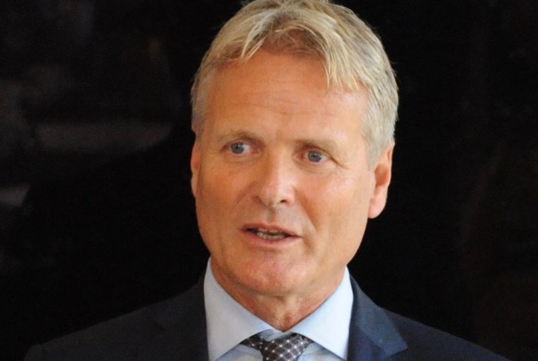 Man in a suit with a light blue shirt and a patterned tie, speaking confidently against a dark background.