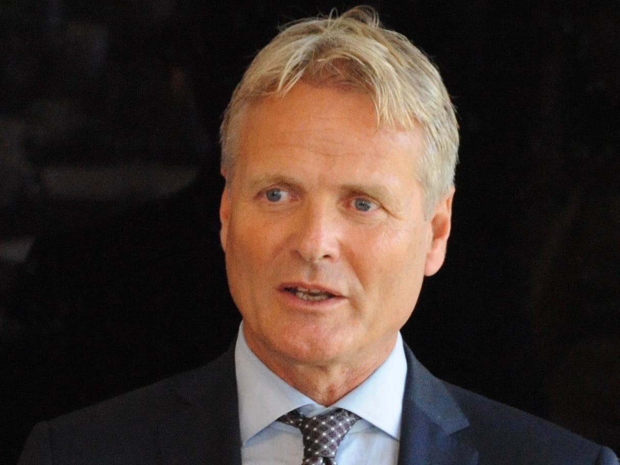 Man in a suit with a light blue shirt and a patterned tie, speaking confidently against a dark background.