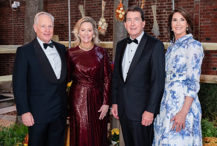 Four adults in formal wear standing together outdoors with decorative plants and lights in the background.