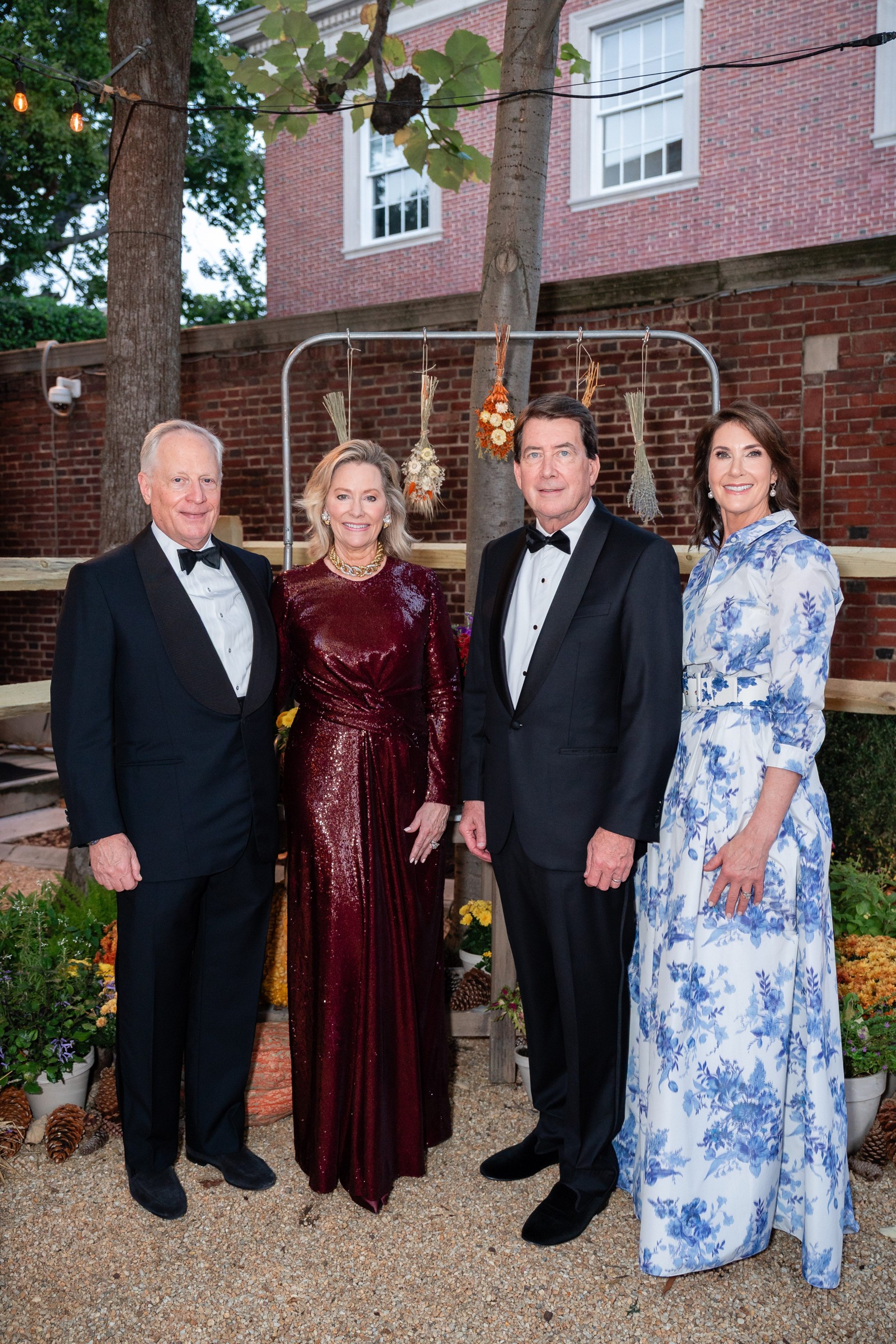 Four adults in formal wear standing together outdoors with decorative plants and lights in the background.